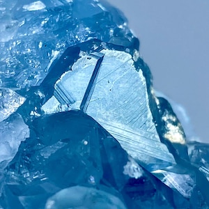 May include: A cluster of translucent light blue crystals featuring sharp, angular facets and geometric edges. The mineral structure displays varying shades of blue with internal light reflections and clear, defined crystalline planes throughout.
