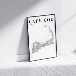 Puede incluir: Impresión enmarcada en negro con un mapa detallado de Cape Cod, Massachusetts, sobre un fondo blanco. El texto "CAPE COD" se muestra en la parte superior. La impresión está apoyada contra una pared blanca sobre un suelo de madera.