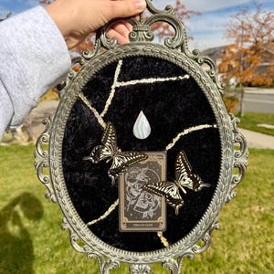May include: An oval, ornate, silver-toned frame holds a black velvet background with gold-colored crackle lines. Two butterflies with black and white wings and a tarot card are affixed to the velvet. A teardrop-shaped stone is centered.