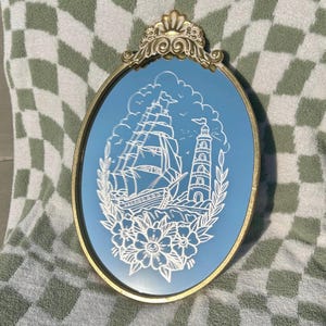 以下が含まれることがあります： 金色のフレームと青色の内側を持つ楕円形の鏡。鏡には、帆船、灯台、花の白い航海デザインが施されています。フレームの上部には装飾的な金のアクセントがあります。