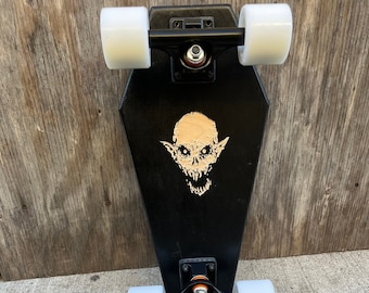 May include: Black coffin-shaped skateboard with a skull graphic and white wheels. The trucks and hardware are black. The skateboard is designed for street or park riding. The deck measures approximately 76 cm in length.