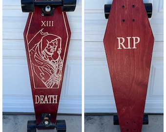 May include: A red wooden skateboard shaped like a coffin with black trucks and wheels. The top of the board has a white graphic of a skeleton holding a scythe with the words "XIII" and "DEATH" on the board. The bottom of the board has the word "RIP" written on it.