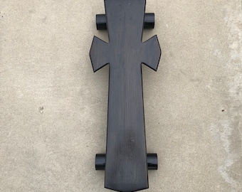 May include: A black wooden skateboard with a cross-shaped design. The skateboard has black wheels and is on a gray concrete surface.