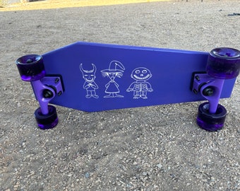 May include: A purple skateboard with white cartoon characters on the deck. The characters are a devil, a witch, and a skeleton. The skateboard has purple wheels.