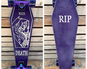 May include: Purple skateboard deck shaped like a coffin with a Grim Reaper graphic and the words "XIII" and "DEATH". The deck has black trucks and purple wheels.