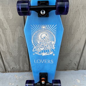 May include: A blue skateboard with black trucks and purple wheels. The deck is shaped like a coffin and features a white graphic of two skeletons holding a heart with the word "LOVERS" printed below.