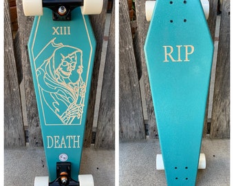 May include: A teal blue skateboard shaped like a coffin with black trucks and white wheels. The top of the board has a gold graphic of a skeleton holding a scythe with the words "XIII" and "DEATH". The bottom of the board has the word "RIP" in gold.