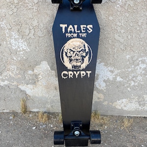 May include: Black coffin-shaped skateboard with black wheels and trucks. The board features the text "TALES FROM THE CRYPT" above a circular graphic of a ghoulish face. The board is set against a textured, light gray wall.