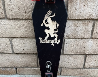 May include: Black skateboard with red wheels and a black and white illustration of Krampus, a mythical creature, on the deck. The deck is shaped like a coffin and has the word "Krampus" written on it.