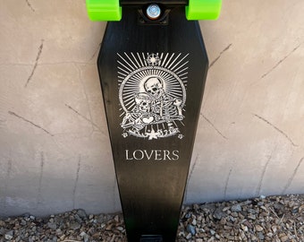 May include: Black longboard skateboard with green wheels and a white graphic of two skeletons holding a heart with the word "LOVERS" printed below.