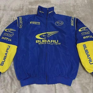 Może przedstawiać: Niebiesko-żółta kurtka Subaru World Rally Team. Kurtka posiada logo Subaru, napis "SUBARU WORLD RALLY TEAM" oraz logotypy sponsorów, takie jak "PIRELLI", "MOTUL" i "Sparco". Rękawy są żółte z niebieskimi mankietami.