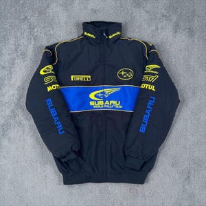 May include: Black Subaru World Rally Team jacket with blue and yellow accents. Features the Subaru logo, Pirelli branding, and sponsor logos. The jacket has a high collar and a full-length zipper.