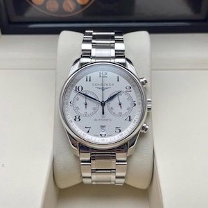 May include: A silver Longines automatic watch with a white face, featuring black hands and numerals. The watch has a stainless steel band and a chronograph function. The watch is displayed on a white cushion.