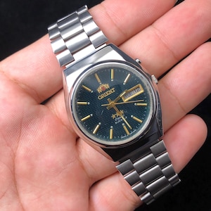 Può includere: Un orologio Orient argentato con quadrante verde scuro e cinturino argentato. Il quadrante mostra lancette e indicatori dorati, con il giorno e la data visibili. L'orologio è di stile classico.