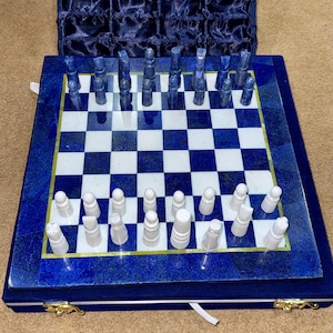 Op de afbeelding: Een schaakspel van blauw en wit marmer met witte stukken op een blauw bord. Het schaakbord zit in een blauwe fluwelen doos met gouden accenten.
