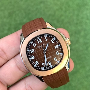May include: A brown Seiko automatic wristwatch with a brown dial and a brown rubber strap. The watch has a gold-colored bezel and a date display at the 3 o'clock position. The watch face has white numerals and hands.