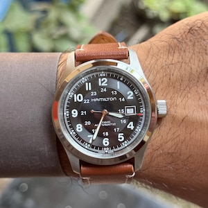 Puede incluir: Un reloj Hamilton Khaki Automatic con caja de acero inoxidable y correa de cuero marrón. La esfera negra del reloj tiene números y agujas blancas. El reloj tiene una pantalla de fecha a las 3 en punto.