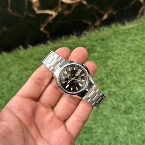 May include: A vintage wristwatch with a silver-tone metal band and a black watch face. The watch face features gold-tone hour markers and hands. The watch is held in a hand against a green background.