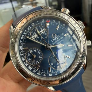 Puede incluir: Reloj automático Omega Speedmaster azul con bisel plateado y correa de caucho azul. La esfera azul tiene múltiples subesferas y una pantalla de fecha. El bisel presenta una escala taquimétrica.