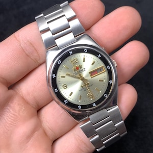 May include: Silver-toned Orient watch with a light gold face, black bezel, and silver band. The watch face displays the Orient logo, day and date, and numeric hour markers. The watch band is made of linked metal segments.