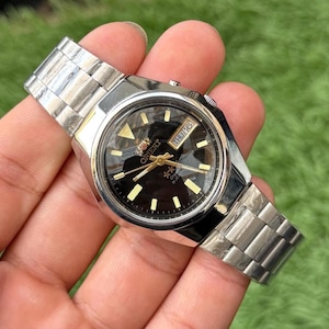 May include: A silver-tone stainless steel wristwatch with a round black dial, gold-tone hour markers, and gold-tone hands. The watch features a day-date window at the three o'clock position displaying 'THU 16' and includes a metal link bracelet.