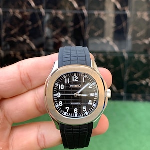 May include: A Seiko automatic wristwatch with a black dial and a black rubber strap. The watch has a silver-colored case and a gold-colored bezel. The watch face displays the time with white numerals and hands. The watch is held by a hand.