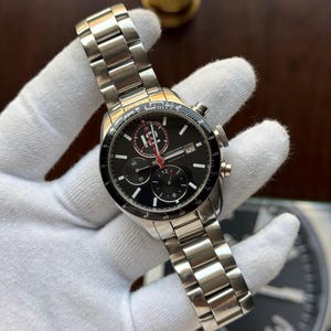 May include: A silver-tone stainless steel Longines automatic chronograph watch with a black dial, three sub-dials, a date window at the three o'clock position, a tachymeter bezel, and a metal link bracelet with a deployment clasp closure.
