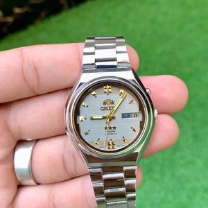 May include: Silver Orient watch with a light grey face, gold hands, and a date display. The watch has a silver strap and a round face with gold accents. The watch face displays the Orient logo and the text "crystal 2 jewels".
