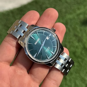 Puede incluir: Un reloj de pulsera plateado con esfera verde, con ventana de fecha a las 3. El reloj tiene una correa de acero inoxidable y una esfera redonda. El reloj se sostiene en una mano sobre un fondo verde.