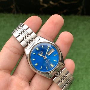 May include: A vintage Seiko automatic wristwatch with a bright blue dial. The watch has a silver-tone stainless steel strap and case. The dial shows the day and date, with "MON 18" visible. The watch has luminous hands and markers.