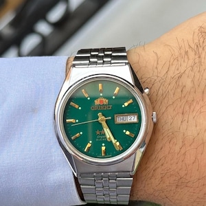 Puede incluir: Un reloj de pulsera Orient con caja redonda de acero inoxidable plateado y brazalete de eslabones a juego. El reloj presenta una esfera verde vibrante, índices y manecillas doradas, y una ventana de día y fecha que muestra THU 27.