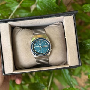 May include: A silver wristwatch with a teal face and a date display, presented in a black box with a beige cushion. The watch has a silver strap and a blue bezel. The watch is a classic style.