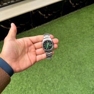 May include: A silver wristwatch with a green dial and a silver band. The watch has a round face with silver hands and markers. The watch is held in a hand against a green background.