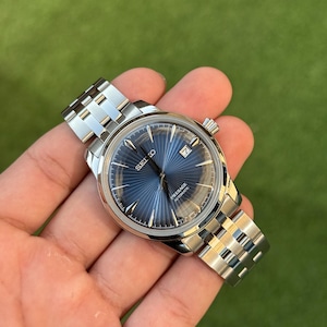 May include: A stainless steel wristwatch with a blue dial. The watch face displays the brand name "SEIKO" and the word "PRESAGE AUTOMATIC". The watch has a silver band and a date display at the 3 o'clock position.