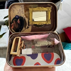Könnte beinhalten: Eine kleine Blechdose mit einem rosa-weißen Herzmuster im Inneren. Die Dose enthält eine goldene Haarklammer, eine kleine Flasche mit einem Korkverschluss und eine klare Glasflasche mit einem weißen Deckel. Auf der Flasche steht "With Love".
