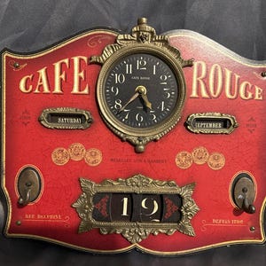 May include: A red wall-mounted calendar clock with a crackled finish, featuring the text CAFE ROUGE. It includes a central analog clock face, two small windows displaying SATURDAY and SEPTEMBER, and a central date display showing 19.