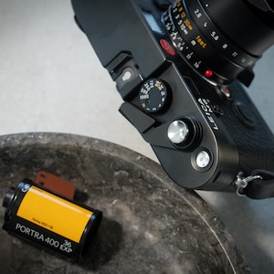 Puede incluir: Una cámara de película negra con una lente plateada y un rollo de película Portra 400 de 36 exposiciones en un contenedor amarillo.