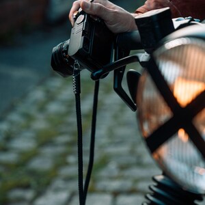 May include: A black camera with a lens is held by a person. The camera is mounted on a motorcycle, with the headlight and handlebars visible. The background is blurred, showing a street and buildings.