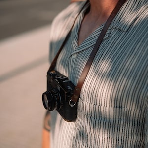 Op de afbeelding: Een zwarte vintage camera met een bruine leren riem hangt om de nek. De camera heeft een lens en diverse knoppen. De riem is bruin. De camera wordt gedragen over een gestreept overhemd.