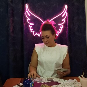 Pode incluir: Uma mulher est&aacute; sentada a uma mesa com uma placa de asa de anjo de n&eacute;on rosa acima de sua cabe&ccedil;a. Ela est&aacute; segurando um baralho de cartas de tar&ocirc; em suas m&atilde;os. H&aacute; um objeto em forma de olho azul e branco na mesa &agrave; sua frente.