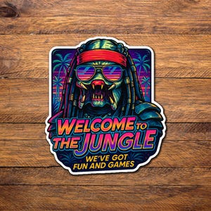 May include: A sticker featuring a stylized Predator character wearing sunglasses and a red headband. The text "WELCOME TO THE JUNGLE" and "WE'VE GOT FUN AND GAMES" are displayed in vibrant colors against a tropical sunset background.