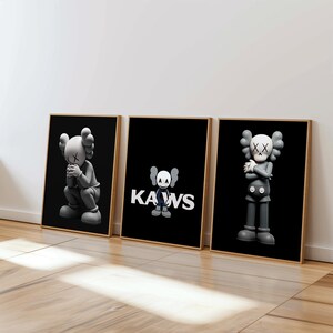 Peut inclure: Trois impressions encadrées représentant des illustrations en noir et blanc du personnage Kaws. L'impression centrale présente le logo Kaws en texte blanc sur fond noir.