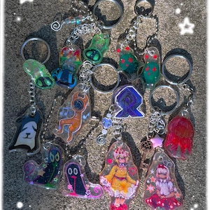 May include: A selection of colourful keyrings featuring diverse designs, such as cartoon characters and abstract shapes. Each keyring has a silver chain and a metal ring. The keyrings are made of clear resin and are suitable for keys or bags.