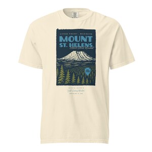 May include: Cream-colored t-shirt featuring a graphic of Mount St. Helens National Volcanic Monument. The design includes the mountain, trees, and text that reads "Mount St. Helens" and "Est. 1982".