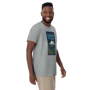 May include: A light gray t-shirt featuring a graphic of Mount St. Helens in a rectangular design. The design includes the text "Mount St. Helens" in blue, a mountain illustration, and trees. The shirt is made of a soft material.