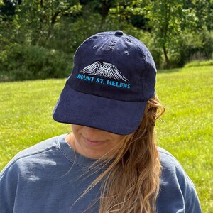May include: Navy blue baseball cap with the text "MOUNT ST. HELENS" embroidered in white and teal. The cap features a mountain graphic and a curved brim. The hat is made of a corduroy material.