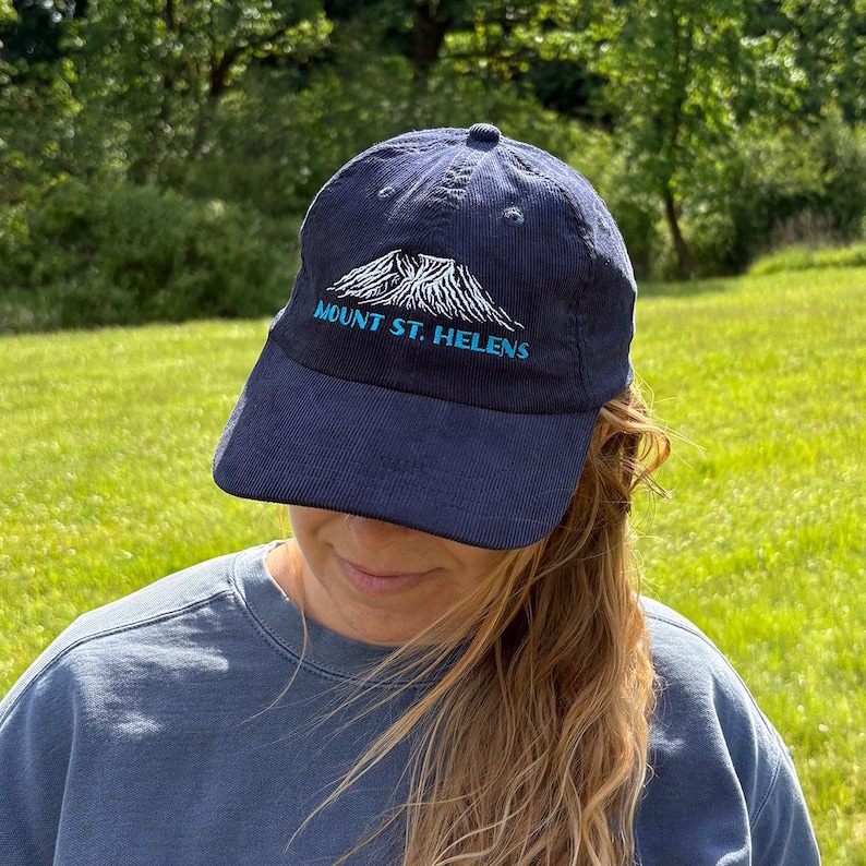 May include: Navy blue baseball cap with the text "MOUNT ST. HELENS" embroidered in white and teal. The cap features a mountain graphic and a curved brim. The hat is made of a corduroy material.