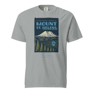 May include: A light gray t-shirt featuring a graphic of Mount St. Helens National Volcanic Monument. The design includes the mountain, trees, and text that reads "Mount St. Helens" and "Gifford Pinchot, Washington".