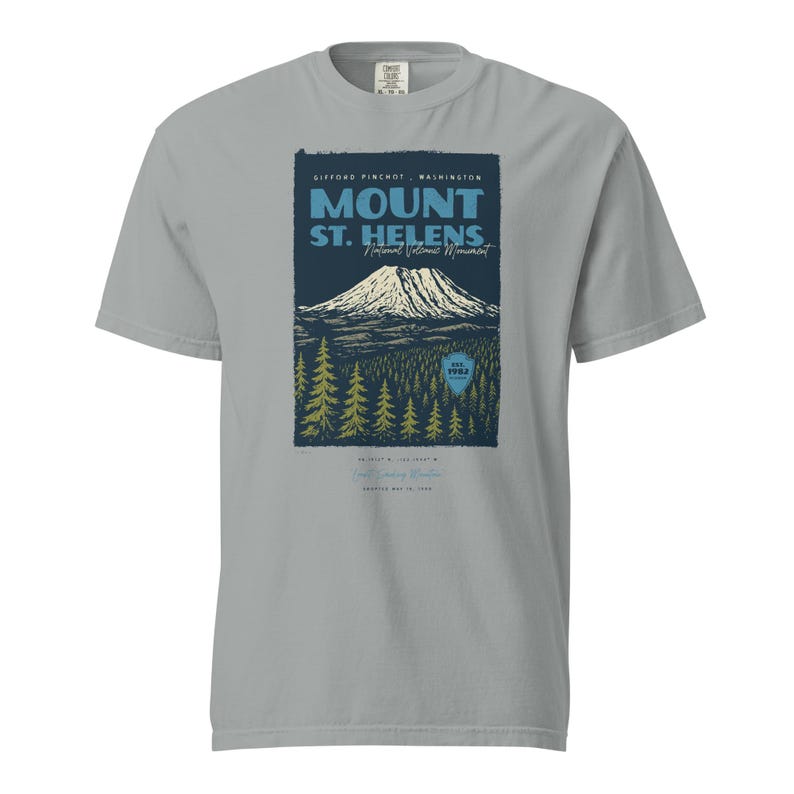 May include: A light gray t-shirt featuring a graphic of Mount St. Helens National Volcanic Monument. The design includes the mountain, trees, and text that reads "Mount St. Helens" and "Gifford Pinchot, Washington".