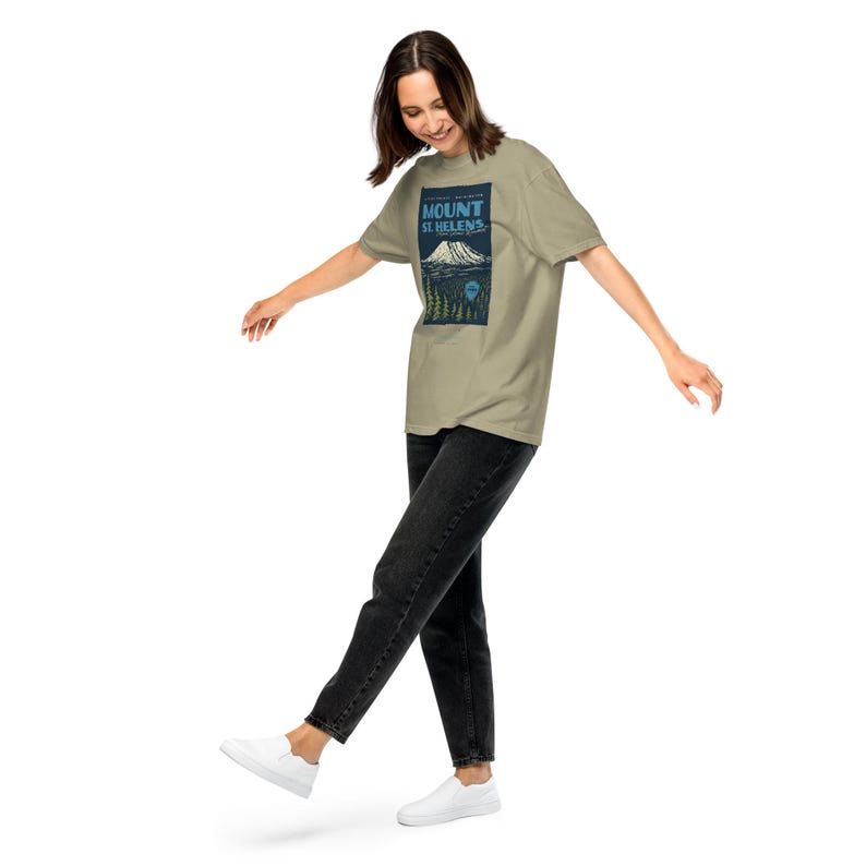 May include: A tan t-shirt featuring a graphic of Mount St. Helens with the text "Mount St. Helens" in blue. The design includes a mountain, trees, and a blue sky. The person is wearing black jeans and white shoes.